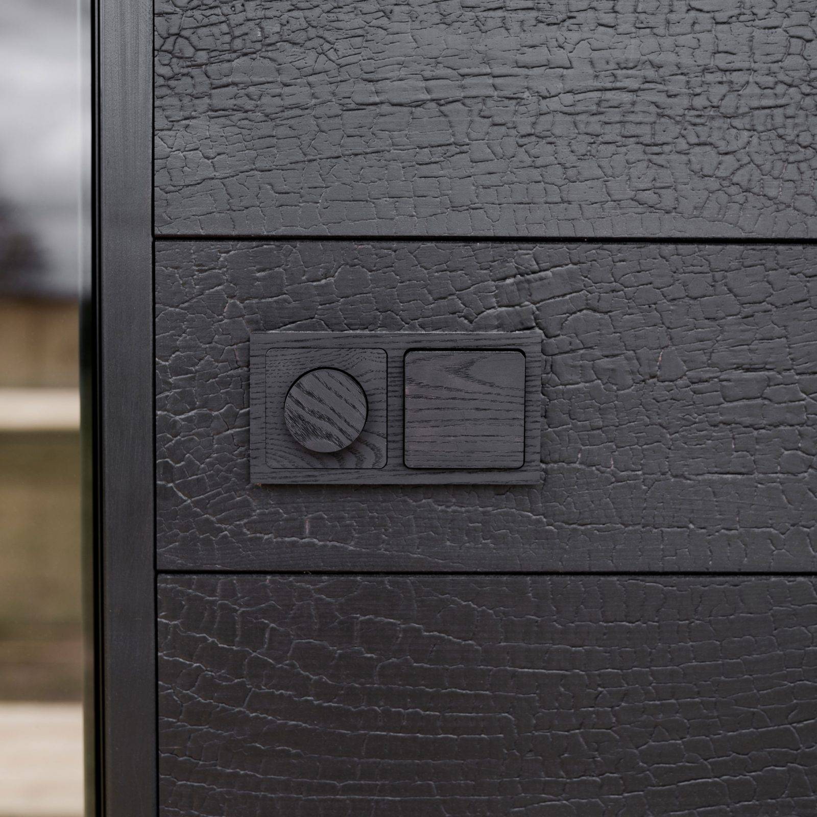 Close-up of a sleek black control panel with textured wood finish, featuring a circular dial and a rectangular button, designed for Ember & Ice outdoor saunas emphasizing modern Scandinavian design and functionality.