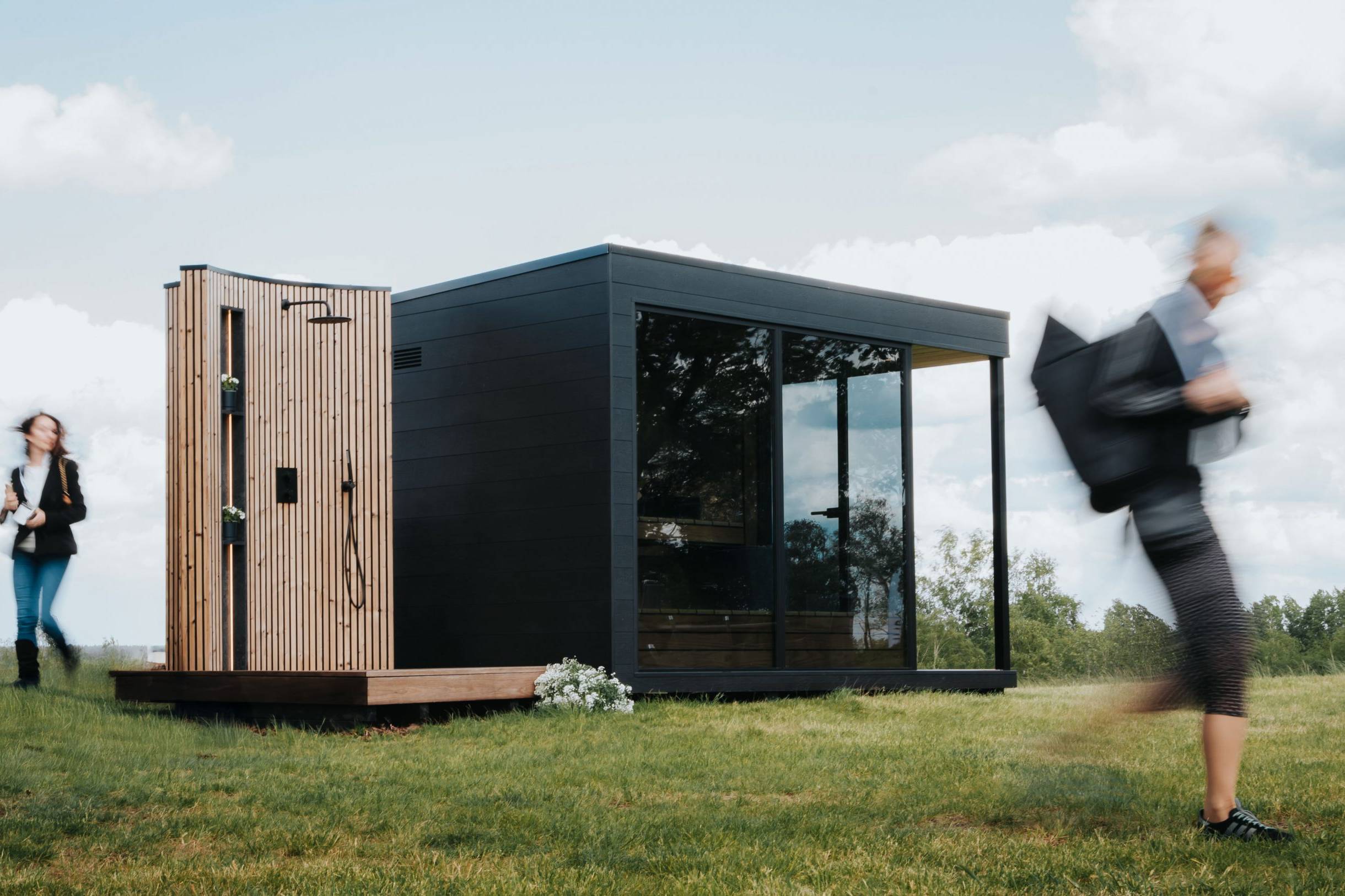 Outdoor sauna with a sleek black design and wooden accents, surrounded by green grass, featuring a person walking by, enhancing the wellness experience offered by Ember and Ice.
